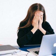 Moteris sėdi prie kompiuterio, šalia daug popierių. Ji užsidengusi veidą delnais